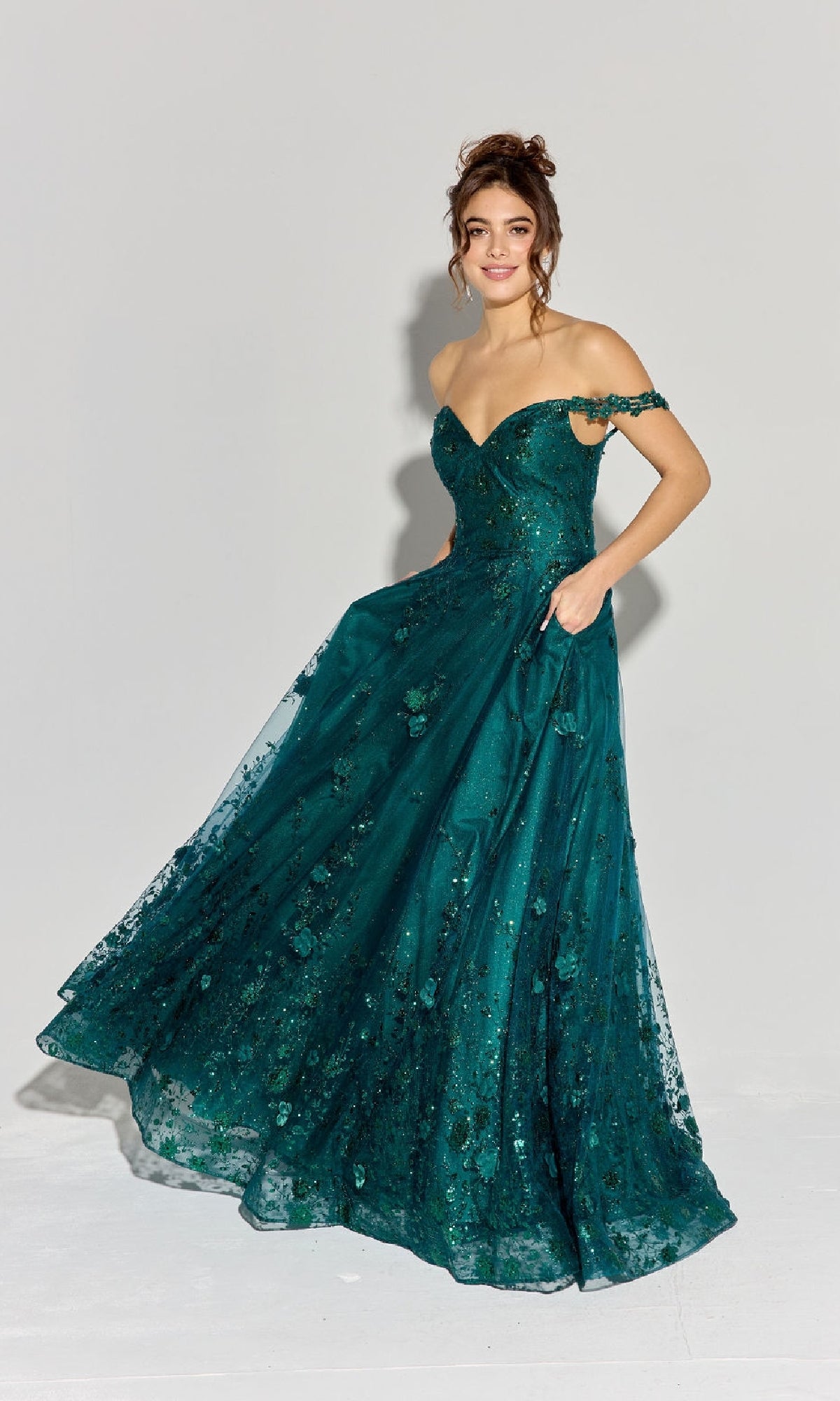 A woman in the Long Glitter-Tuller A-Line Prom Dress Eureka 9980, featuring a teal off-the-shoulder design with floral patterns, stands on a plain background holding part of the skirt.